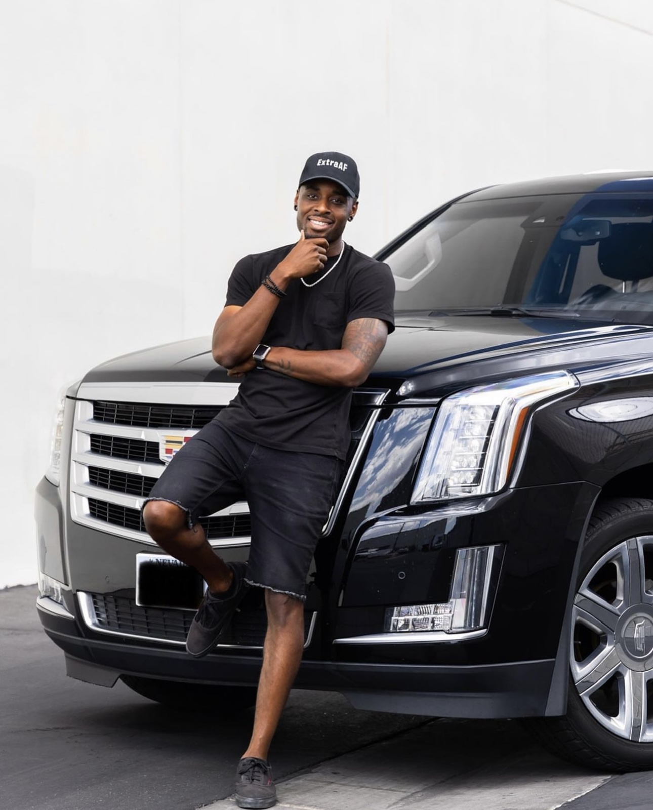 A man in black casual wear leans on a black Cadillac SUV, smiling confidently against a white background.